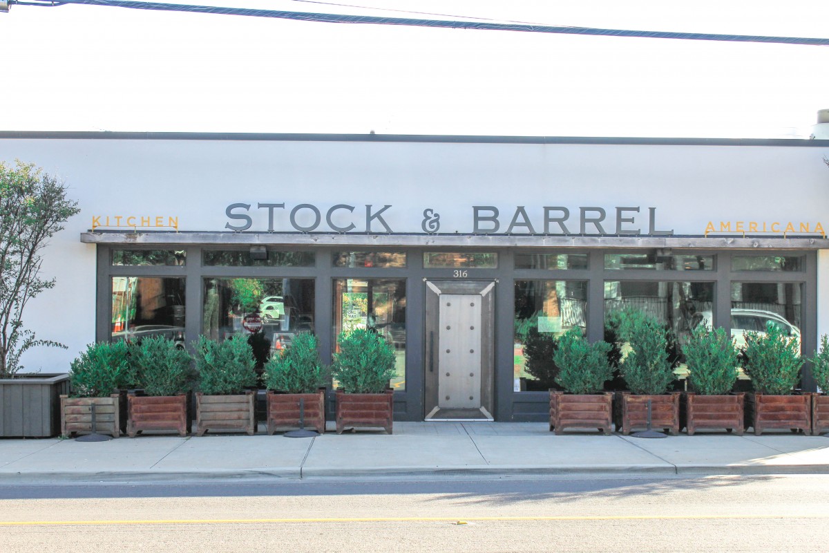 This is the entrance of Stock & Barrel in Bishop Arts District. It's a plain white building with several plants lined outside. 