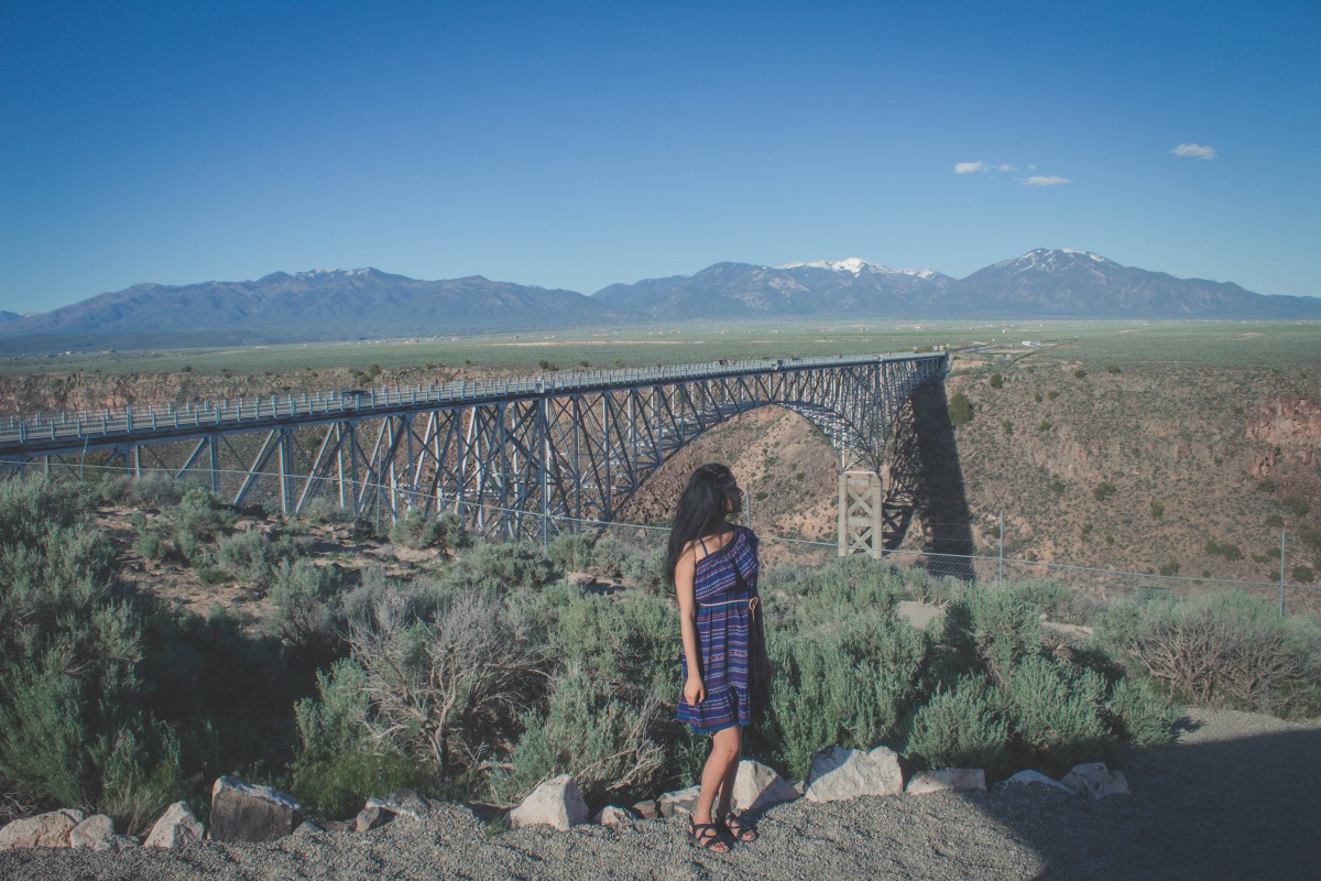 Girl in blue dress standing in front of Rio Grande Gorge Bridge. 