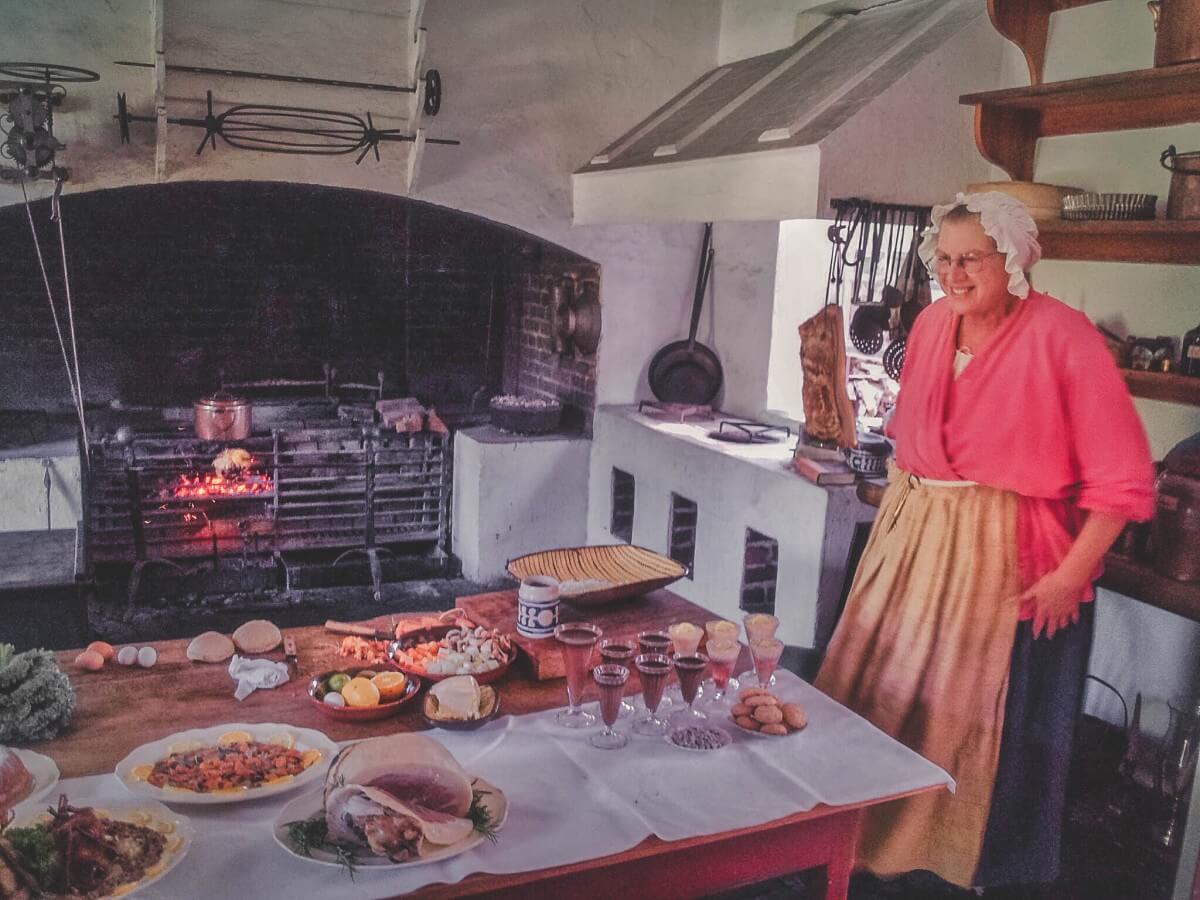 a woman, dressed in period clothing, smiles in front of a full feast of food at one of the cooking demonstrations in Colonial Williamsburg. She's wearing a bonnet and there's a fire in the back heating up food. 