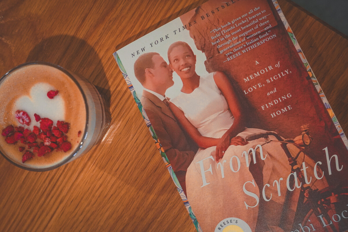 a table at a cafe with a copy of From Scratch beside a cappuccino with heart shaped latte art (to represent books about travel and love)