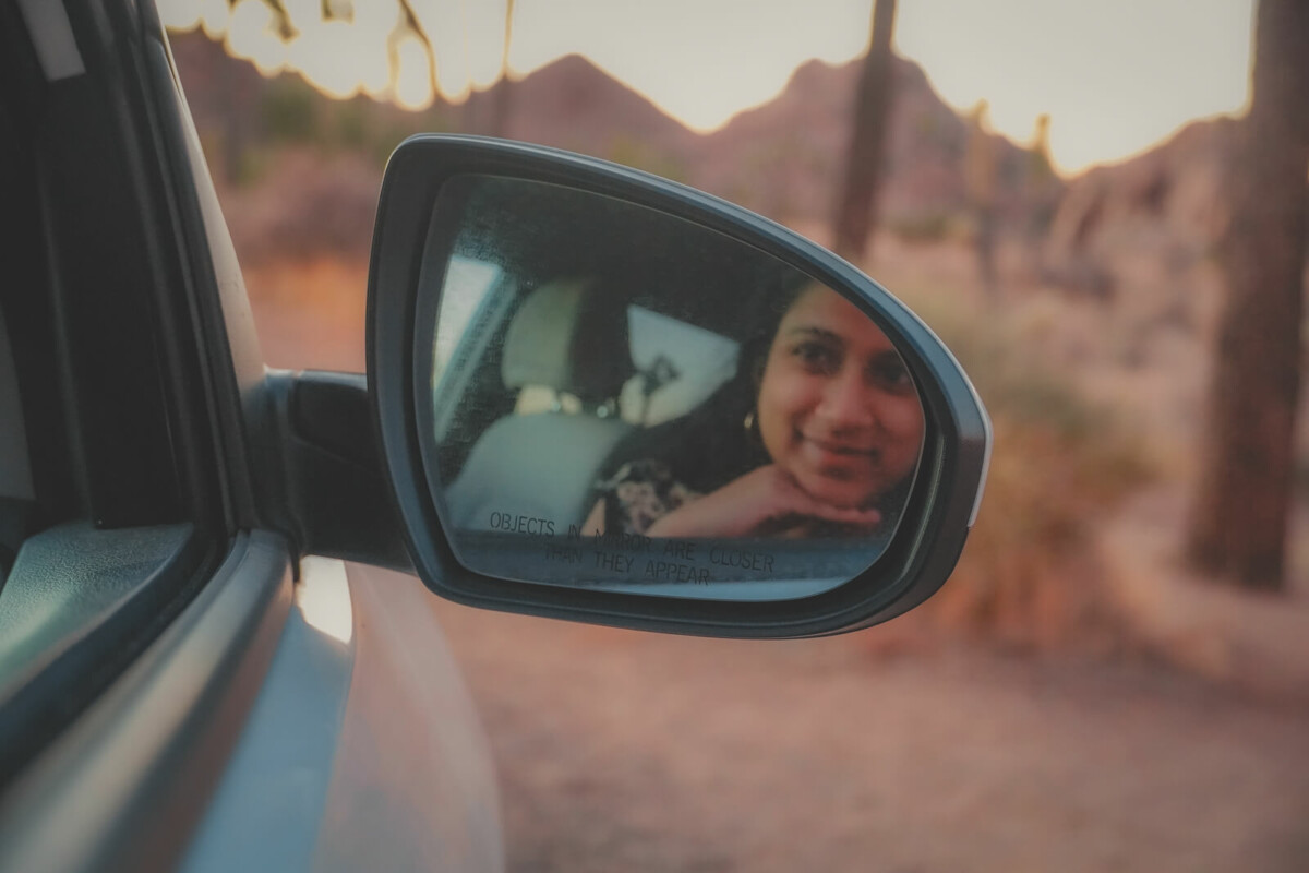 girl smiling at side view mirror of a car with the text objects in the mirror are closer than they appear to represent would you rather road trip questions