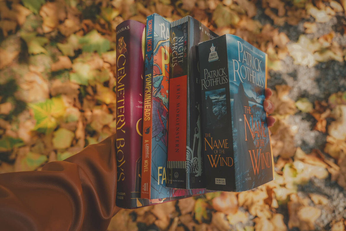 a photo of a sidewalk full of fall leaves and a hand carrying four autumn books, from Pumpkinheads to The Name Of The Wind