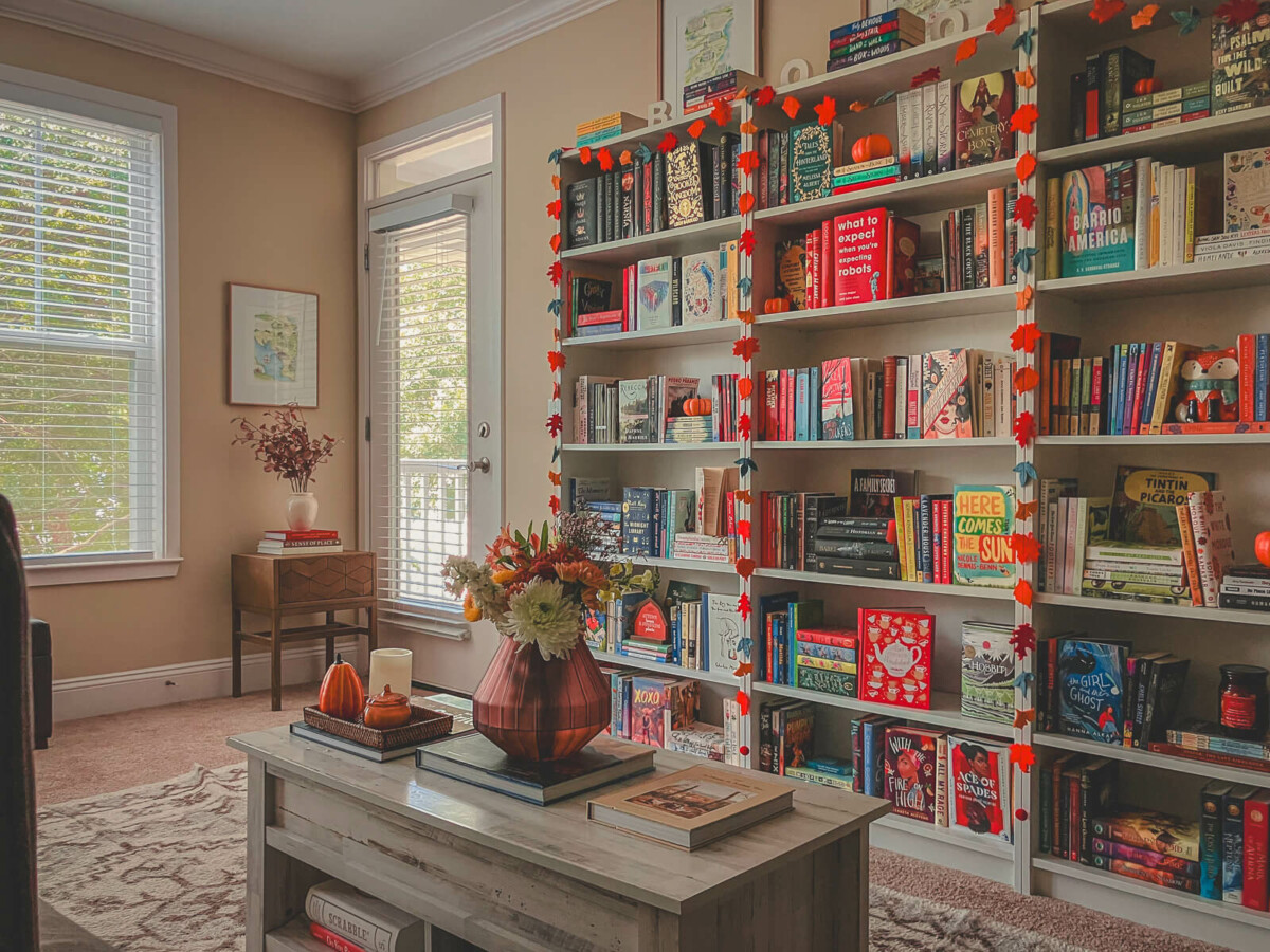 a living room bookshelf, on a sunlit autumn evening, full of cozy autumn books, a draping of fall leaves, pumpkins, and candles