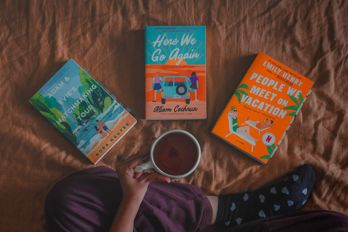 Spicy travel romance books (Adam & Evie's Matchmaking Tour, Here We Go Again, People We Meet On Vacation) spread out in front of a woman holding a mug with a heart at the bottom of the cup. 
