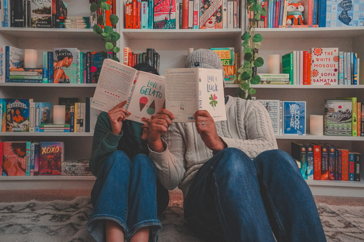 a couple holding up two of the best ya travel romance books, Love & Gelato and another novel in the Love & Other Detours series, in front of a bookshelf filled with travel books